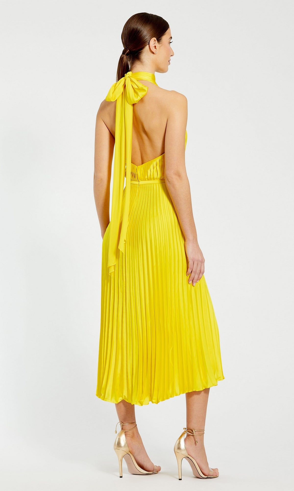 A woman faces away, wearing the Mac Duggal Tea Length Formal Dress 26909—sleeveless, pleated, tea length with a high neck and bow—paired with metallic high-heeled sandals.